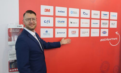 Ein Repa-Mitarbeiter zeigt auf eine Wand mit Firmenlogos.