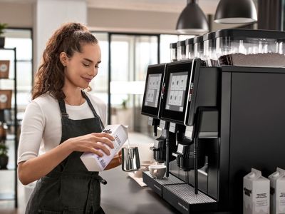 Weibliche Barista gießt vor einem Kaffeevollautomaten Milch in ein Kännchen.