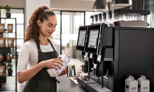 Weibliche Barista gießt vor einem Kaffeevollautomaten Milch in ein Kännchen.