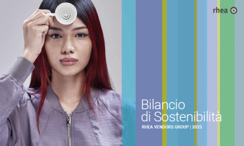 Eine Frau mit langen schwarz-roten Haaren, Nasenring und lilafarbenen Blouson hält eine Espressotasse vor ihrer Stirn. Bilancio di Sostenibilita Rhea Vendors Group 2025
