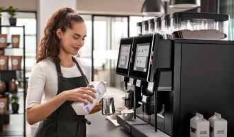 Weibliche Barista gießt vor einem Kaffeevollautomaten Milch in ein Kännchen.