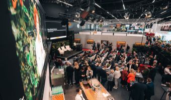 In einer Messehalle sitzen viele Menschen auf Stühlen vor einer Bühne, auf der leere Stühle stehen.